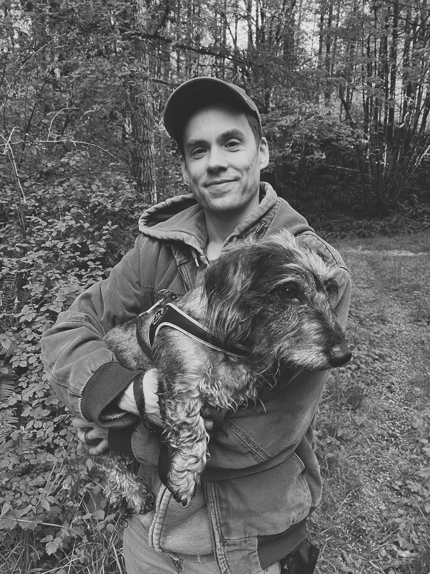 Chris standing in a northwest forest during Spring, while holding Finley (a standard wire-haired dachshund)